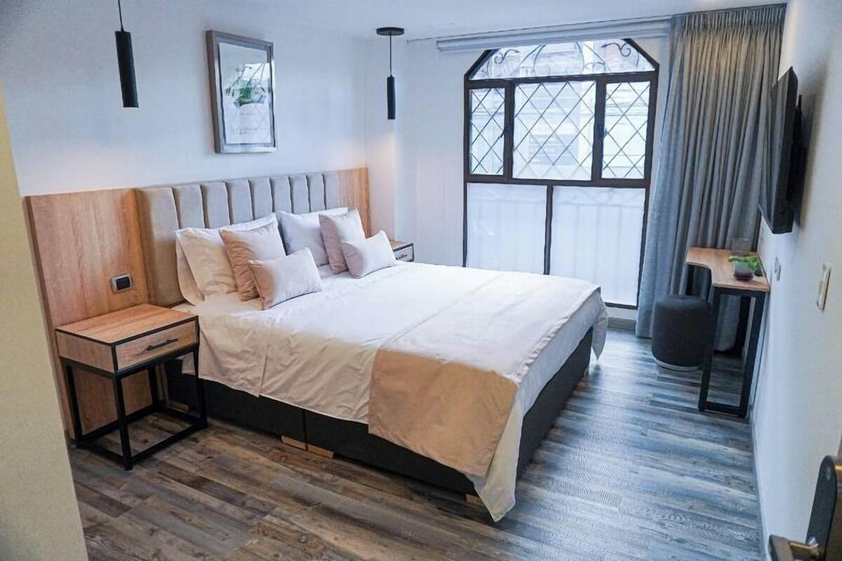 Ein modernes Schlafzimmer mit einem großen Bett, einem Holz-Kopfteil, Nachttischen, einem Fenster mit Vorhängen und einem an der Wand befestigten Fernseher.