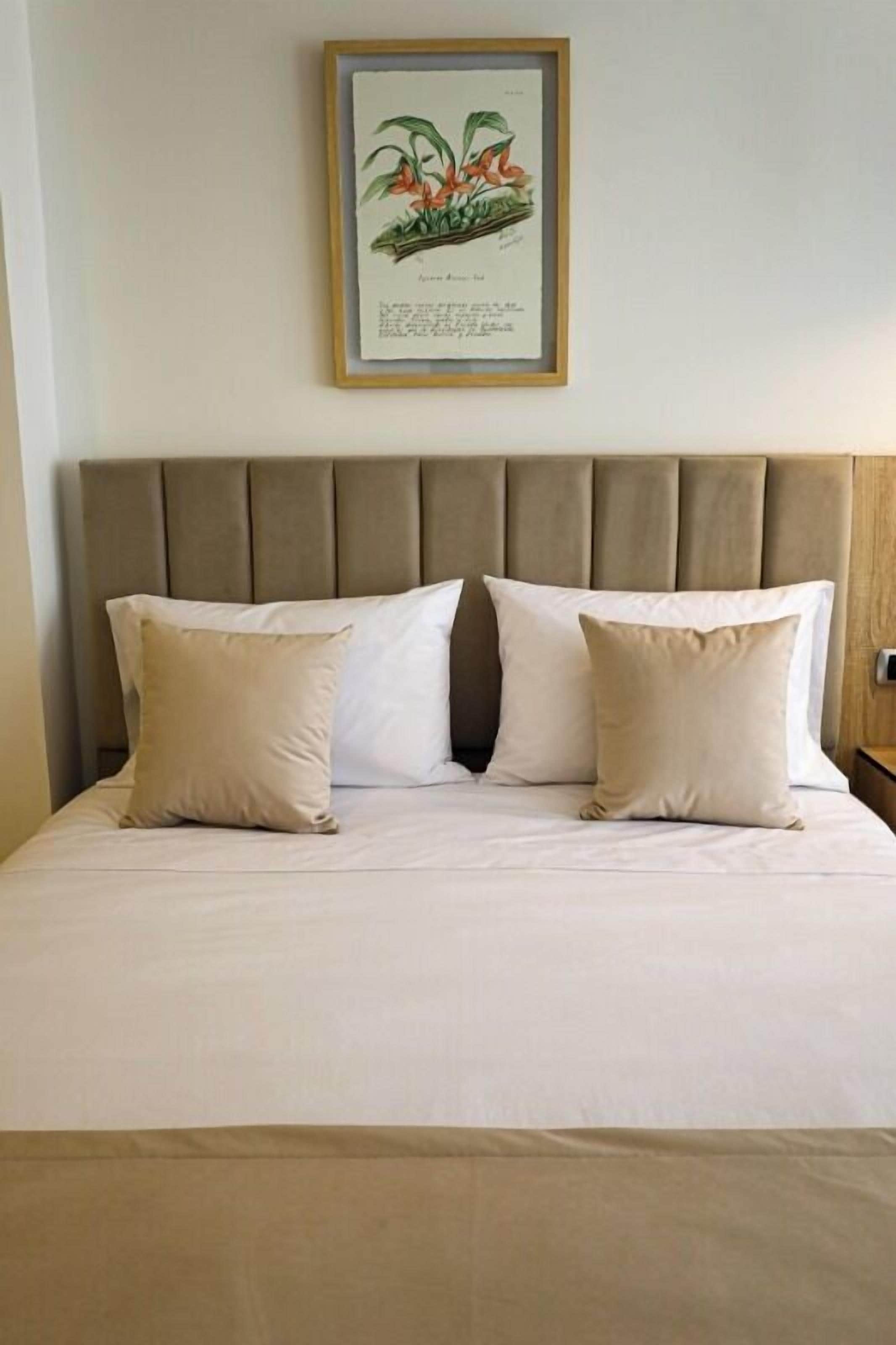 A neatly made bed with a headboard, two pillows, and a framed botanical illustration above it.