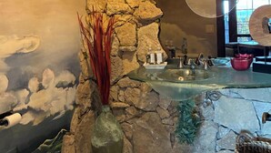 Ein Badezimmer mit Steinwand, einem Glaswaschbecken und einer Vase mit roten Pflanzen.