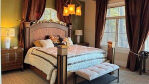 Ein Schlafzimmer mit einem großen Bett, einem Nachttisch, einer Bank und einem Fenster mit Vorhängen.