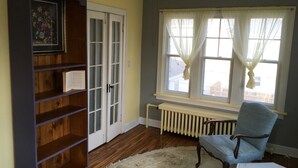 Una habitación con estantería de madera, radiador, ventana con cortinas, una silla y una mesa.