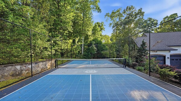 Un court de tennis en plein air, entouré d’une clôture, avec des arbres et un bâtiment en arrière-plan.