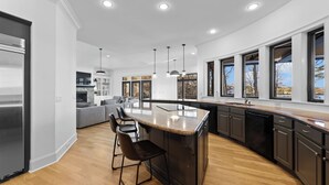 Une cuisine moderne dotée d’un grand îlot central, de meubles de rangement aux couleurs sombres et offrant une vue sur l’extérieur grâce à de grandes fenêtres.