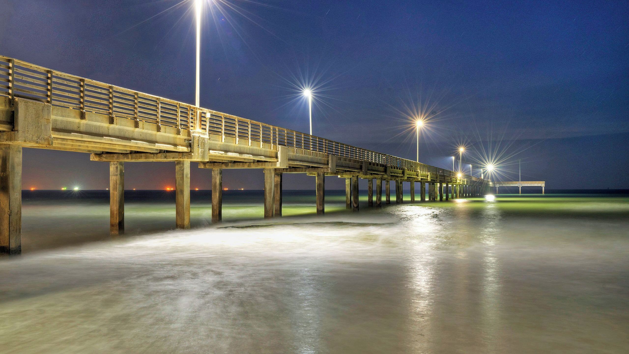 Un muelle que se adentra en el agua, con pasarela y barandillas, bajo un cielo nocturno con iluminación artificial.