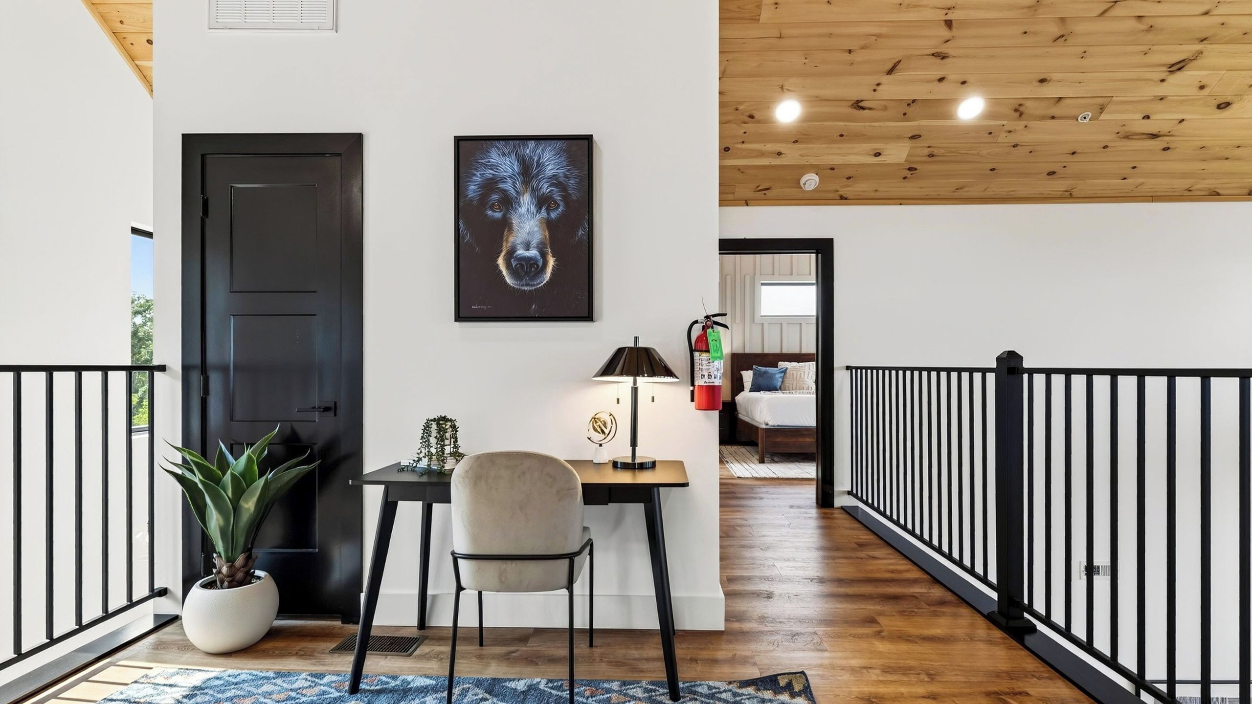 A modern interior with a wooden ceiling, a black door, a framed artwork, a desk with a lamp, a chair, and a potted plant.