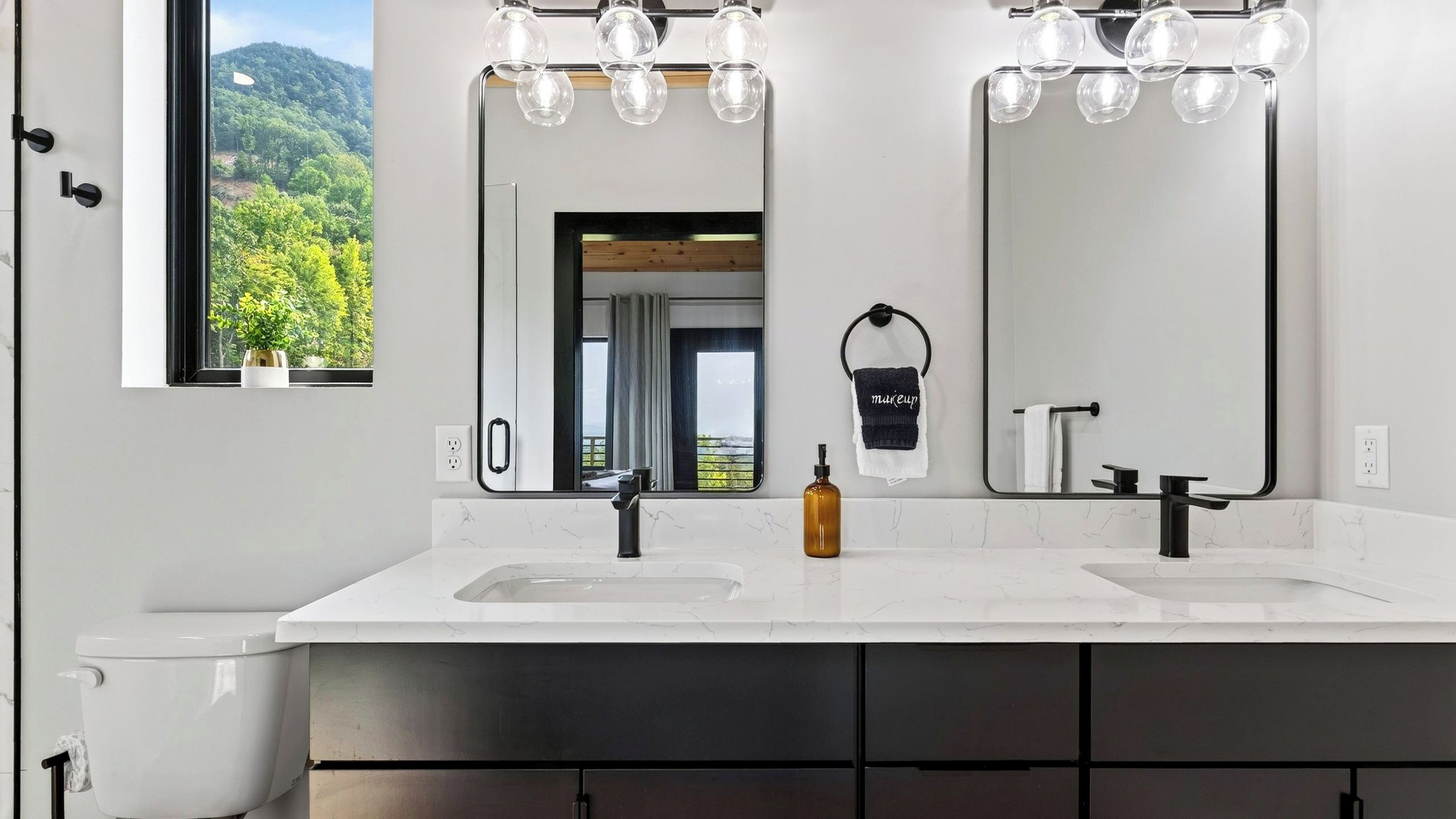 Ein modernes Badezimmer mit zwei Waschbecken, einem großen Spiegel und Blick auf einen Wald durch das Fenster.