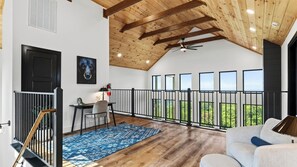 Ein geräumiges Zimmer mit hohen Decken, Holzbalken, einem Balkon mit Geländer, einem Schreibtisch mit Stuhl und einem großen Fenster mit Blick.