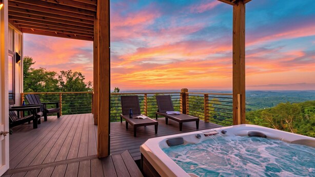 A wooden deck with a hot tub, lounge chairs, and a view of a sunset over a mountainous landscape. - Modern Smoky Mountain Retreat w/ Hot Tub, Game Room & Movie Theater Heavens View by Avantstay (Pigeon Forge)