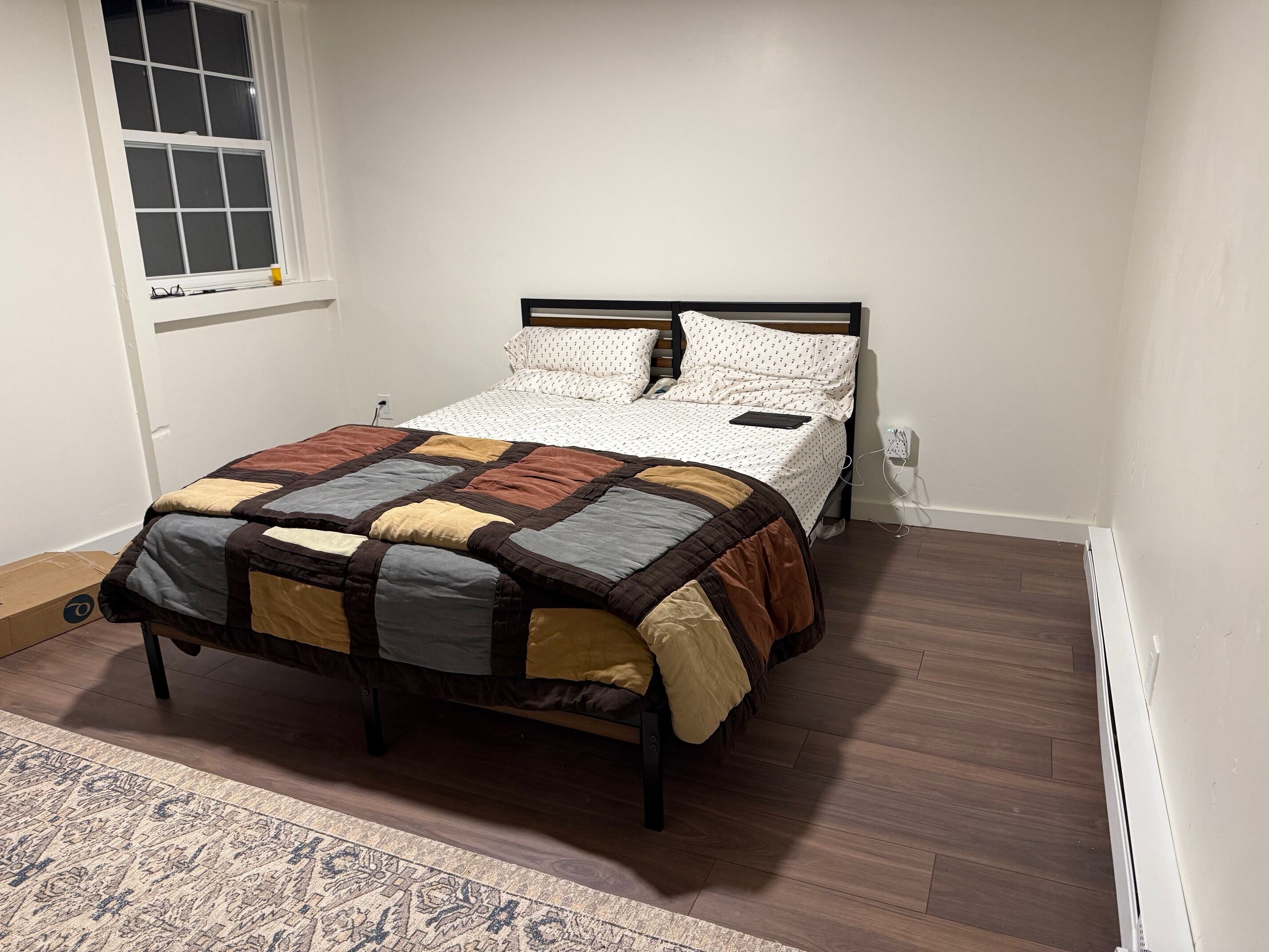 A neatly made bed with a patterned quilt, a bedside table with a lamp, and a window with blinds.