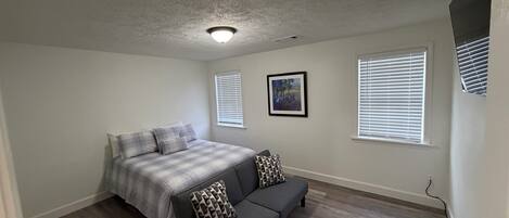 A bedroom with a bed, a gray sofa, two windows with blinds, a ceiling light, and a painting on the wall.