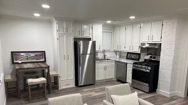 Una cocina moderna con armarios blancos, electrodomésticos de acero inoxidable y una zona de comedor con una mesa de madera y dos sillas.