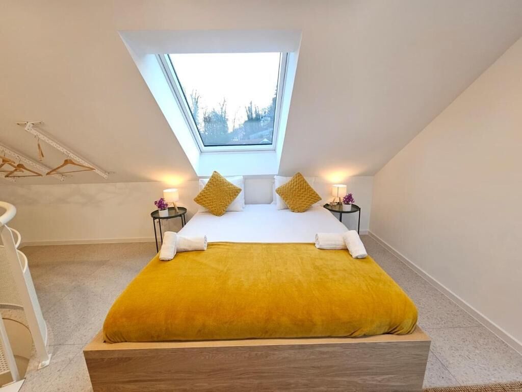 Un dormitorio con tragaluz, una cama con manta amarilla, dos mesitas de noche con lámparas y un cabecero de madera.