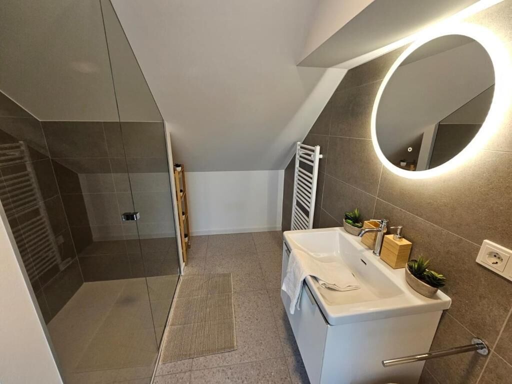 Une salle de bain moderne avec une douche à l’italienne, un lavabo blanc et un miroir doté d’un éclairage circulaire.