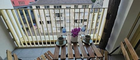 Uma varanda com mesa e cadeiras de madeira, um vaso de planta e vista para prédios e árvores.