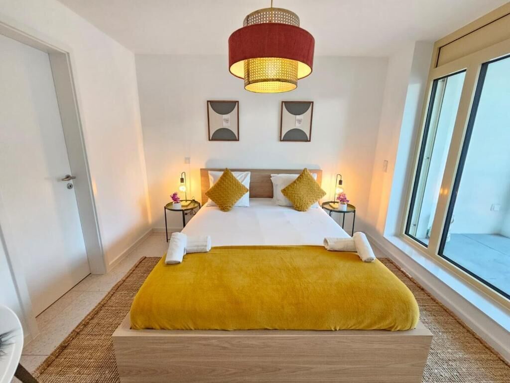 A hotel room with a large bed, two bedside tables, a red pendant light, and a large window.