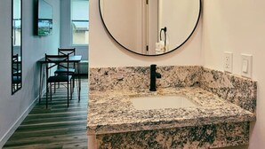 A bathroom with a granite countertop sink, a round mirror, and a wall-mounted light fixture.