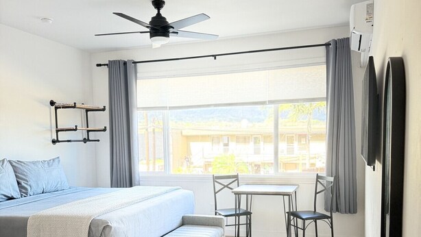 A modern bedroom with a bed, a small sofa, a dining table with chairs, a ceiling fan, and a large window with curtains.