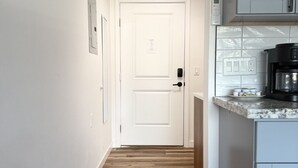 Ein schmaler Flur mit Holzboden, der zu einer weißen Tür mit einem Schild führt. Es gibt eine Küchenzeile mit Arbeitsplatte und Schränken sowie eine Deckenleuchte.
