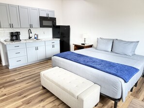 Een kleine slaapkamer met een bed, nachtkastje, bureau, magnetron, koelkast, koffiezetapparaat en wastafel.