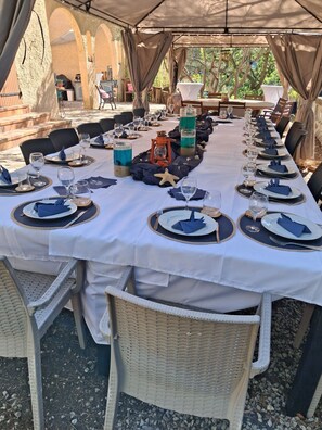 Un área de comedor al aire libre con mesas dispuestas para una comida, rodeada de sillas de mimbre y cubierta por una marquesina.