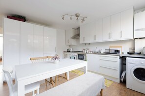 Moderni keittiö, jossa on valkoiset kaapit, ruokapöytä tuoleineen, penkki, pesukone ja kattovalaisin.