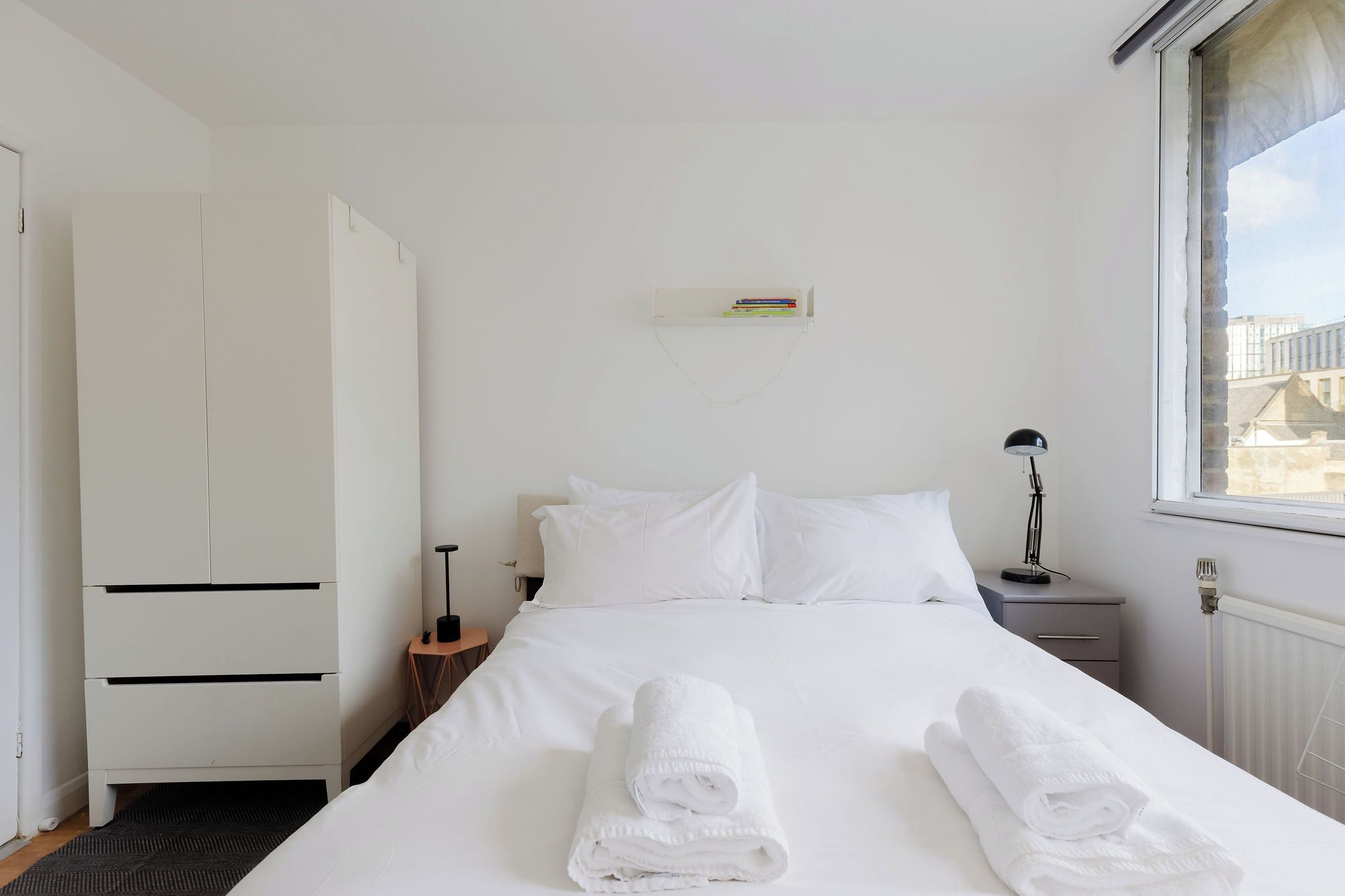 Ett hotellrum med en säng, en garderob, ett nattduksbord, en lampa och utsikt över staden genom fönstret.