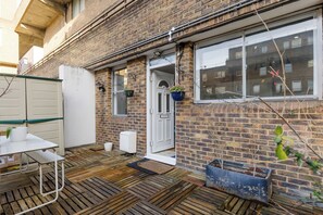 A brick house with a wooden deck, a white door, and a window with a hanging plant. - Brilliantly Located 2BDR Flat in Barbican (Greater London)