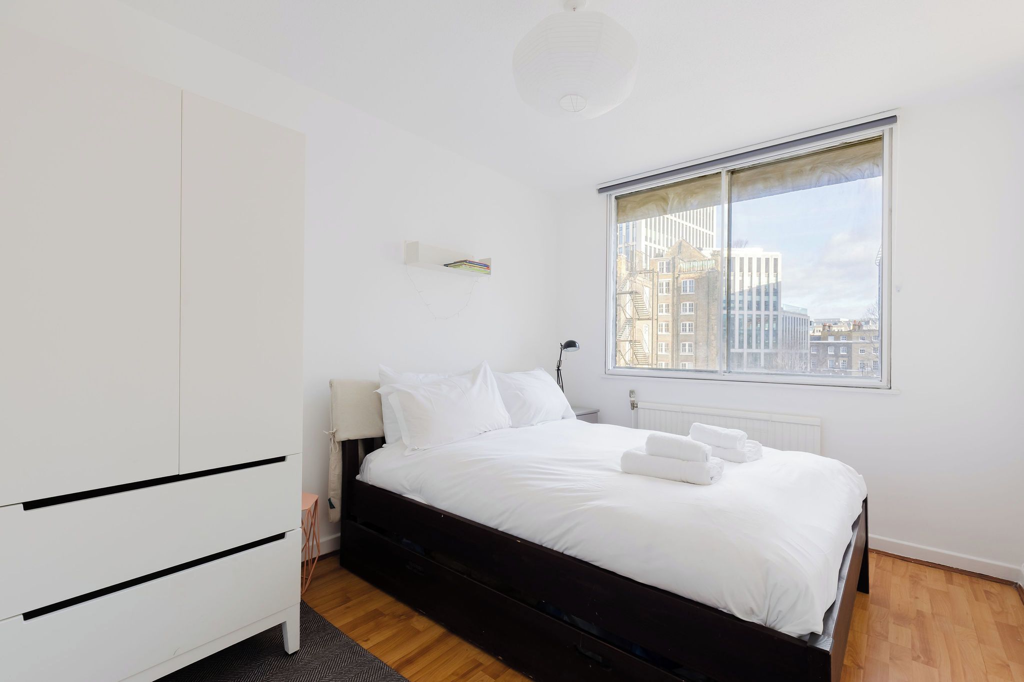 Ein modernes Schlafzimmer mit Bett, Kleiderschrank, Fenster mit Stadtblick und Deckenleuchte.