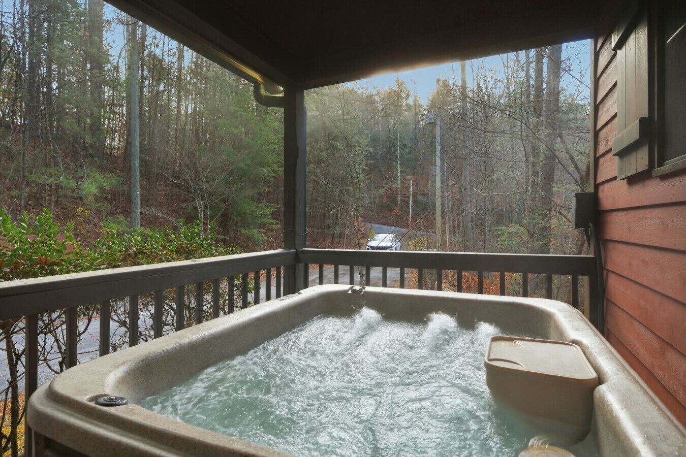 Un bain à remous sur une terrasse en bois, avec vue sur une forêt et un véhicule en arrière-plan.