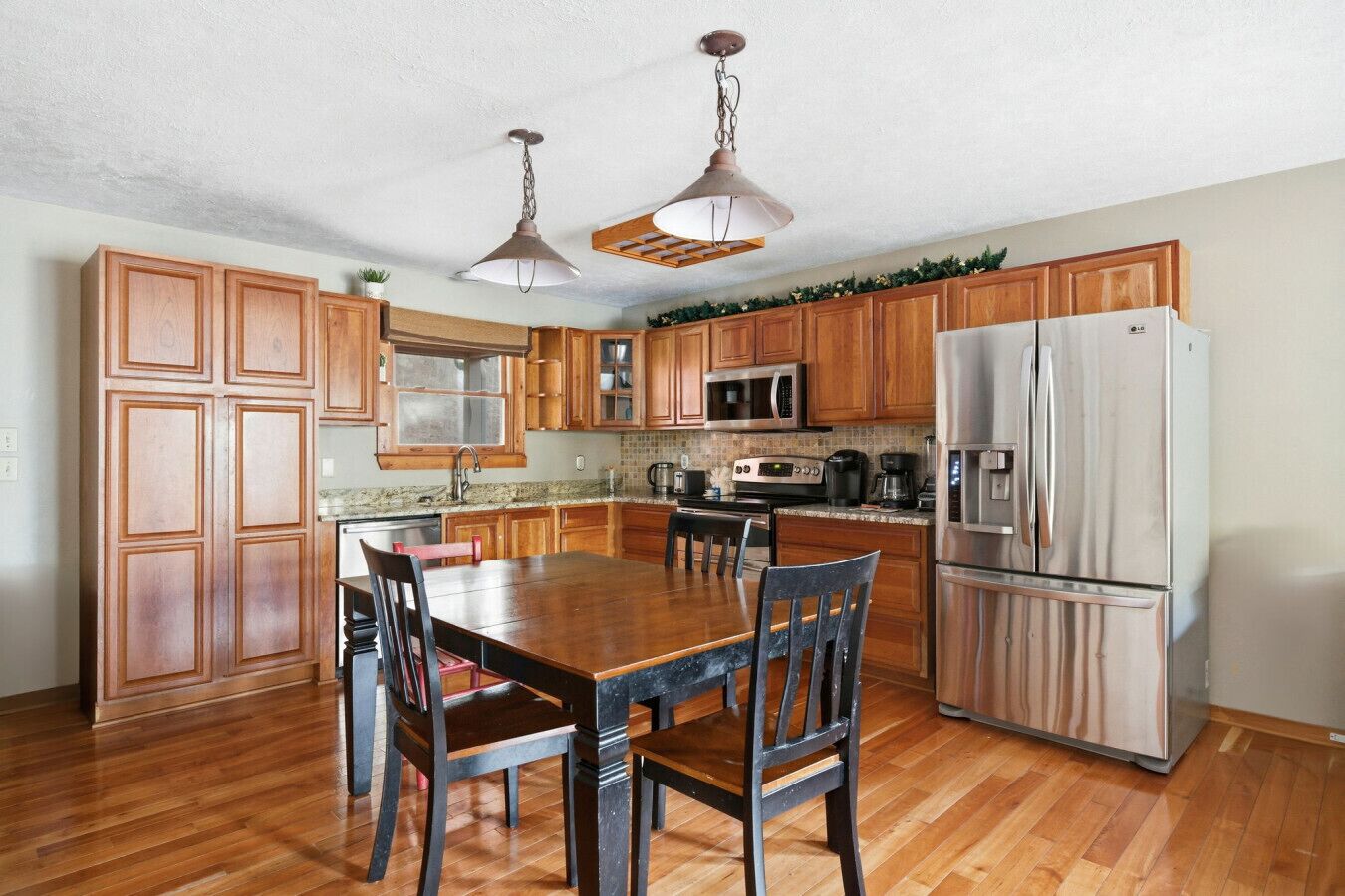 A kitchen with wooden cabinets, a stainless steel refrigerator, a dining table with chairs, and pendant lights.