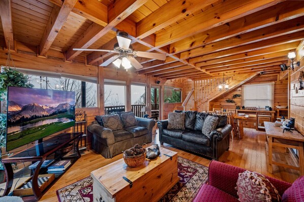 A cozy cabin interior with a wooden ceiling, a ceiling fan, a large TV, two leather sofas, a wooden dining table, and a red chair. - Smokies Mountain Charm: Views, Hot Tub, Game Room! (Sevierville)