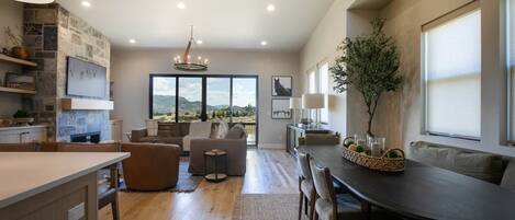 A spacious living area with a large window, a dining table, and a kitchen area with a stone wall.