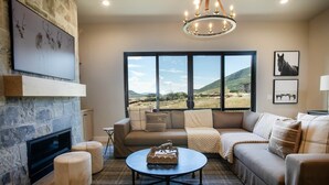 A living room with a stone fireplace, a large window with a view of the countryside, a sofa set, and a round coffee table. - 4BR Deer Valley East | Hot Tub | Shuttle + Views (Wasatch County)
