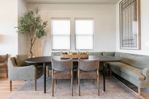 Una sala de estar moderna con una mesa redonda, dos sillas grises, un sofá gris, un ventanal grande y una obra de arte que abarca toda la pared.