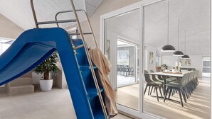 Ein modernes Interieur mit einer blauen Rutsche, einem Essbereich mit langem Tisch und Stühlen sowie einem großen Fenster mit Blick ins Freie.