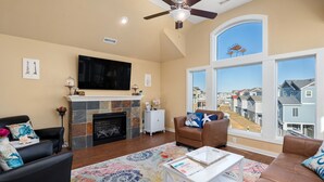 A living room with a fireplace, a ceiling fan, a large TV, and a view of the neighborhood through large windows. - 8Min Walk2Beach,Hot tub, GameRM, Family (Corolla)