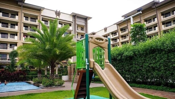 Ein Spielplatz mit Rutsche und Klettergerüst, umgeben von einem Schwimmbad und üppigem Grün, mit Wohngebäuden im Hintergrund.