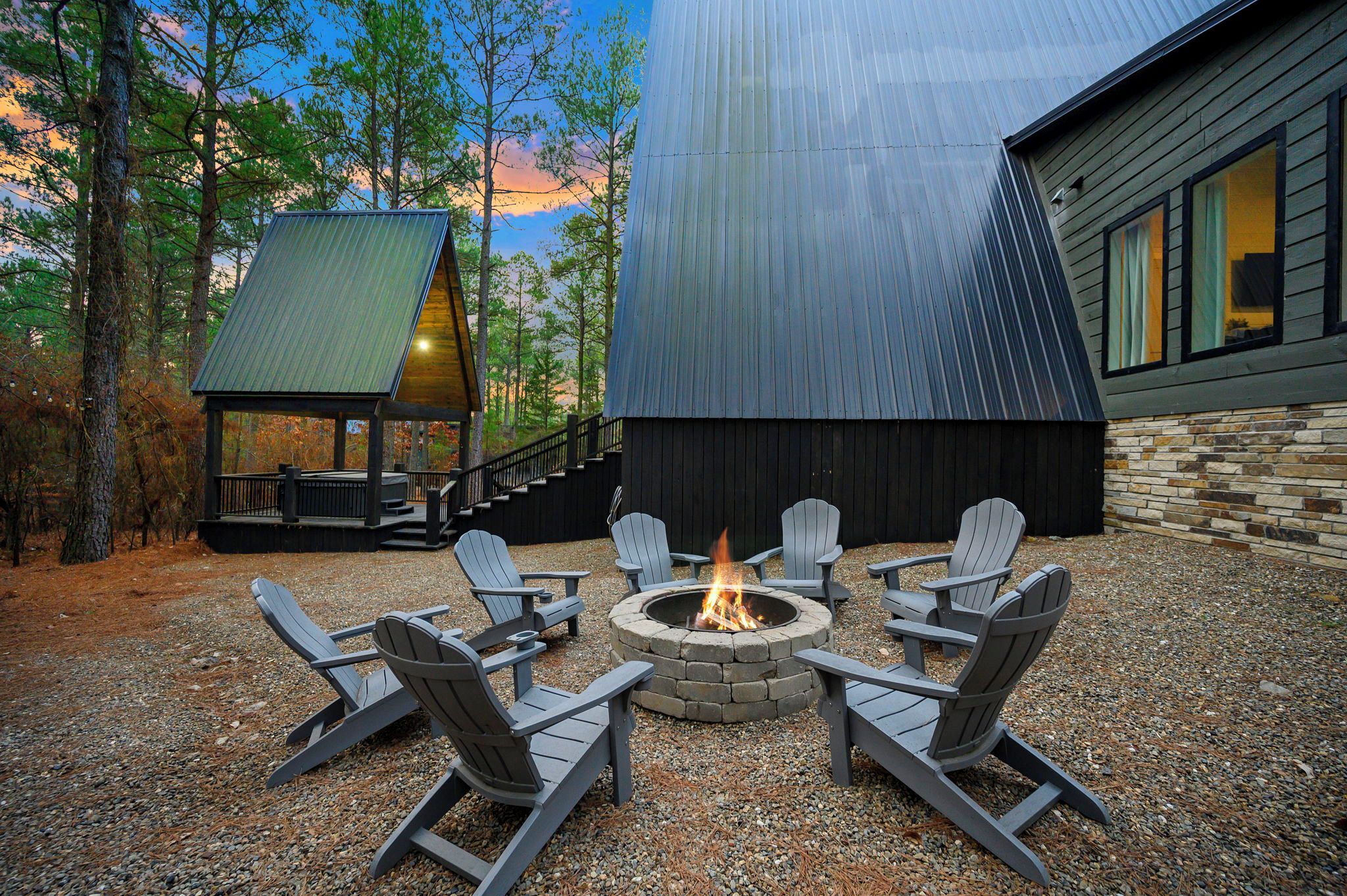 A fire pit surrounded by Adirondack chairs in a gravel area with a wooden deck and a stone wall.