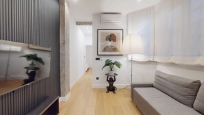 Ein modernes Wohnzimmer mit grauem Sofa, einem kleinen runden Tisch mit Pflanze, einem gerahmten Kunstwerk an der Wand und einem großen Fenster mit Vorhängen.