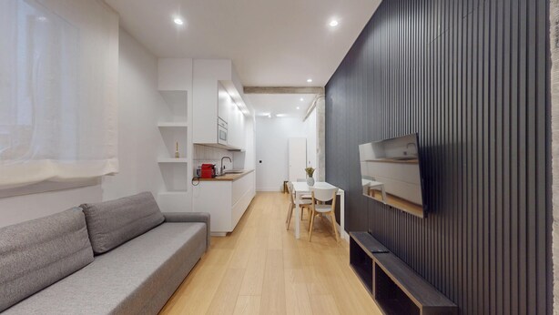 Ein modernes Wohnzimmer mit einer grauen Couch, einer Essecke mit weißem Tisch und Stühlen, einer Küche mit weißen Schränken und einem Fernseher, der an einer dunklen Wand befestigt ist.