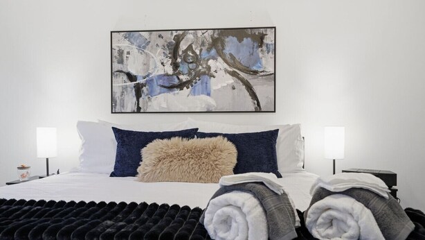 Een net opgemaakt bed met een zacht kussen, twee bedlampen en een groot abstract schilderij aan de muur.