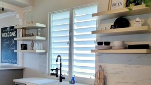 A kitchen with white cabinets, a white countertop, and a window with blinds. There are shelves above the counter with various items, and a chalkboard with "WELCOME" written on it. - Beachy Oasis/Solar Power/Pool/Vibe 4 all ages/Big Groups 4 Family Gatherings (Manatí)