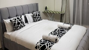 A neatly made bed with a grey upholstered headboard and white bedding, a modern pendant light, and a small table with a green and black patterned tablecloth. - Modern 1-bedroom Near Airport & Nairobi National Park (Nairobi)