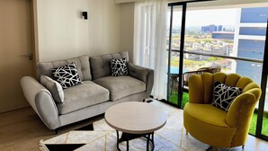 A living room with a grey sofa, a yellow armchair, a round coffee table, and a large window with a city view. - Modern 1-bedroom Near Airport & Nairobi National Park (Nairobi)