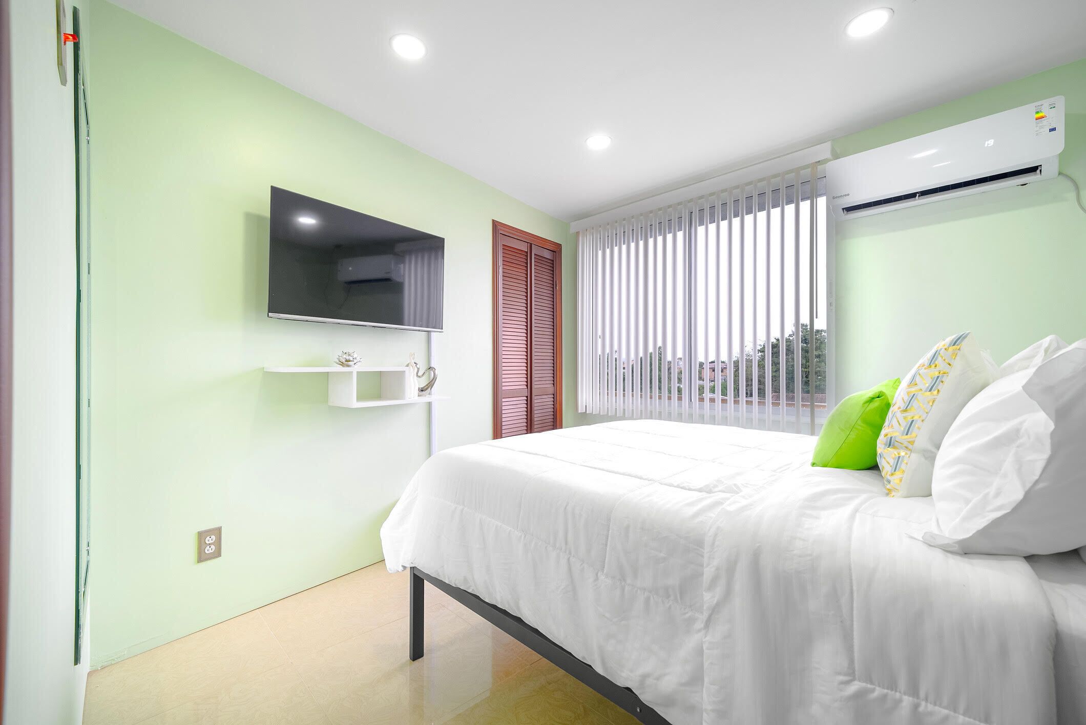 Un dormitorio moderno con cama, televisor montado, equipo de aire acondicionado y ventana con persianas.
