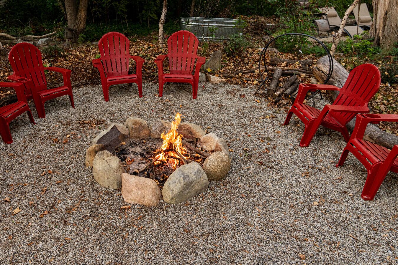 A fire pit surrounded by red Adirondack chairs in a gravel area with trees and logs in the background.