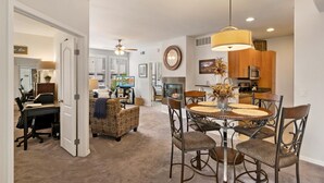 A spacious living room with a fireplace, a dining area with a round table, and a kitchen with wooden cabinets. - Cozy 2-bedroom condo in North Scottsdale with AC, two pools, spas, fitness room (Phoenix)