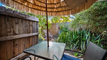 A glass table under a thatched umbrella, a wooden fence, and various plants.