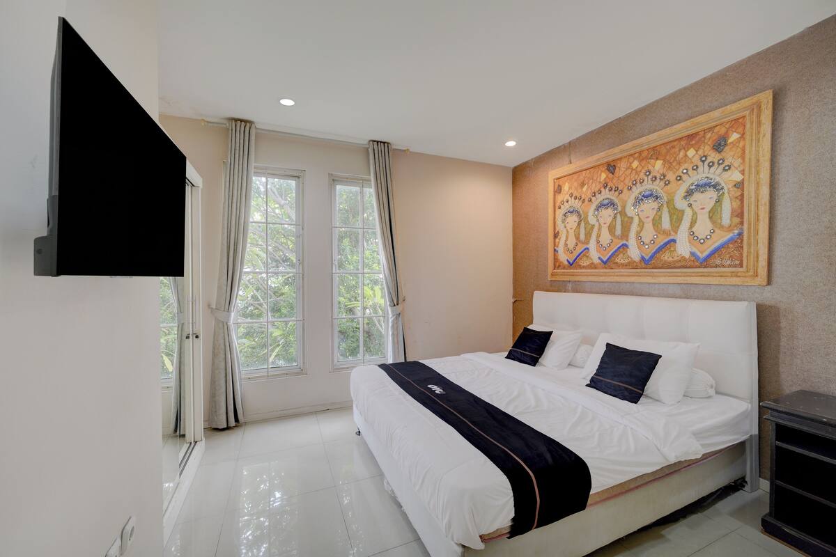 A bedroom with a bed, a painting, a TV, and a window with curtains.