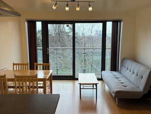 A room with a dining table, chairs, a sofa, and a view through large windows. - Bright 1BDR Apartment with Park Views in Leyton (Greater London)